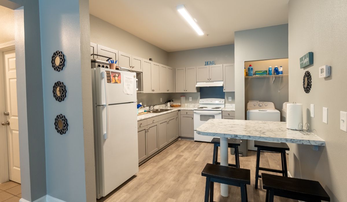 A kitchen with a fridge, gray cabinetry, stove with a hood. The laundry units are attached next to the kitchen and a high chair dining table is attached to the wall opposite the fridge.